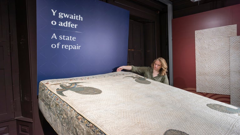 A state bed cover is laid out on display, with its intricate details visible. A property curator stands nearby, examining the bed cover closely.